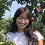 An East Asian woman is smiling while standing in the middle of a plum/apricot orchard.