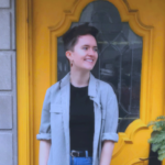 A short-haired Caucasian woman in casual wear is standing in front of a yellow door.