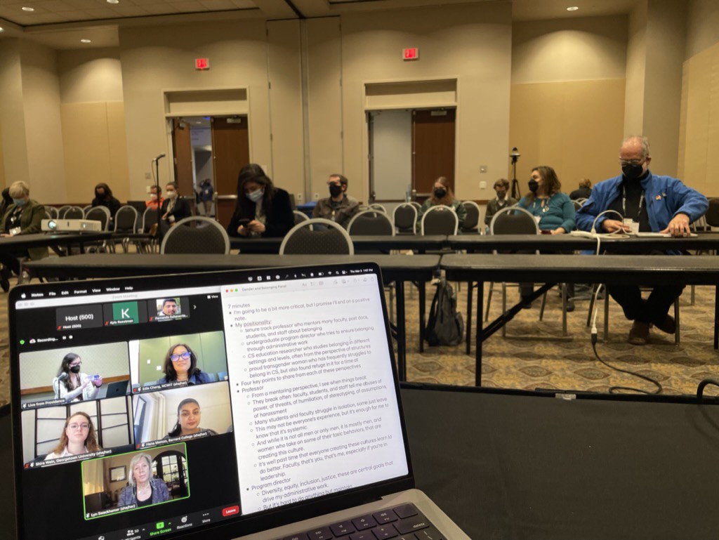 Conference with people on chairs and laptop screen with people talking on it alongside speaker notes.   