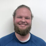 James is smiling in front of a white background. His hair is pulled back in a ponytail and he is wearing a blue shirt.