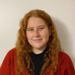 Emily poses in front of a beige wall while wearing a red sweater