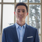 Connor poses in front of a window while wearing a blue suit jacket and light blue button up shirt.