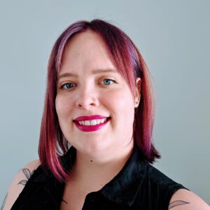 Dr. Morrison Smith rocks purple hair in front of a greyish blue background. She is wearing a black button down sleeveless shirt and hints of tattoos are showing.