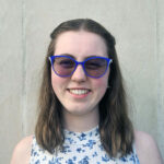 Leah poses in front of a white background. She has shoulder length brown hair and is wearing blue cat eye glasses