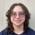 Soren poses in front of a white background. He is wearing a navy t shirt and has dark brown curly hair to his shoulders. He is wearing round glasses.