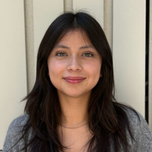 Juliette poses in front of a beige building while wearing a grey sweater