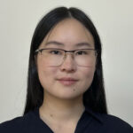 Sydney is wearing half rim glasses and a dark blue button up while posing in front of a blank background