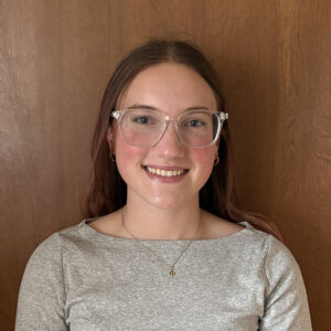 Gwen is a brunette Caucasian woman with clear rimmed glasses. She is posing in front of a brown door while wearing a grey knit blouse.