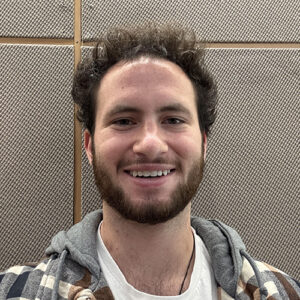 Royce is a caucasian male with brown hair and a beard. He is smiling in front of a brown wall while wearing a plaid hooded jacket and a white t shirt