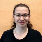 Madeline poses in front of a wooden wall. She is a caucasian female with brown curly hair and is wearing glasses and a black t shirt