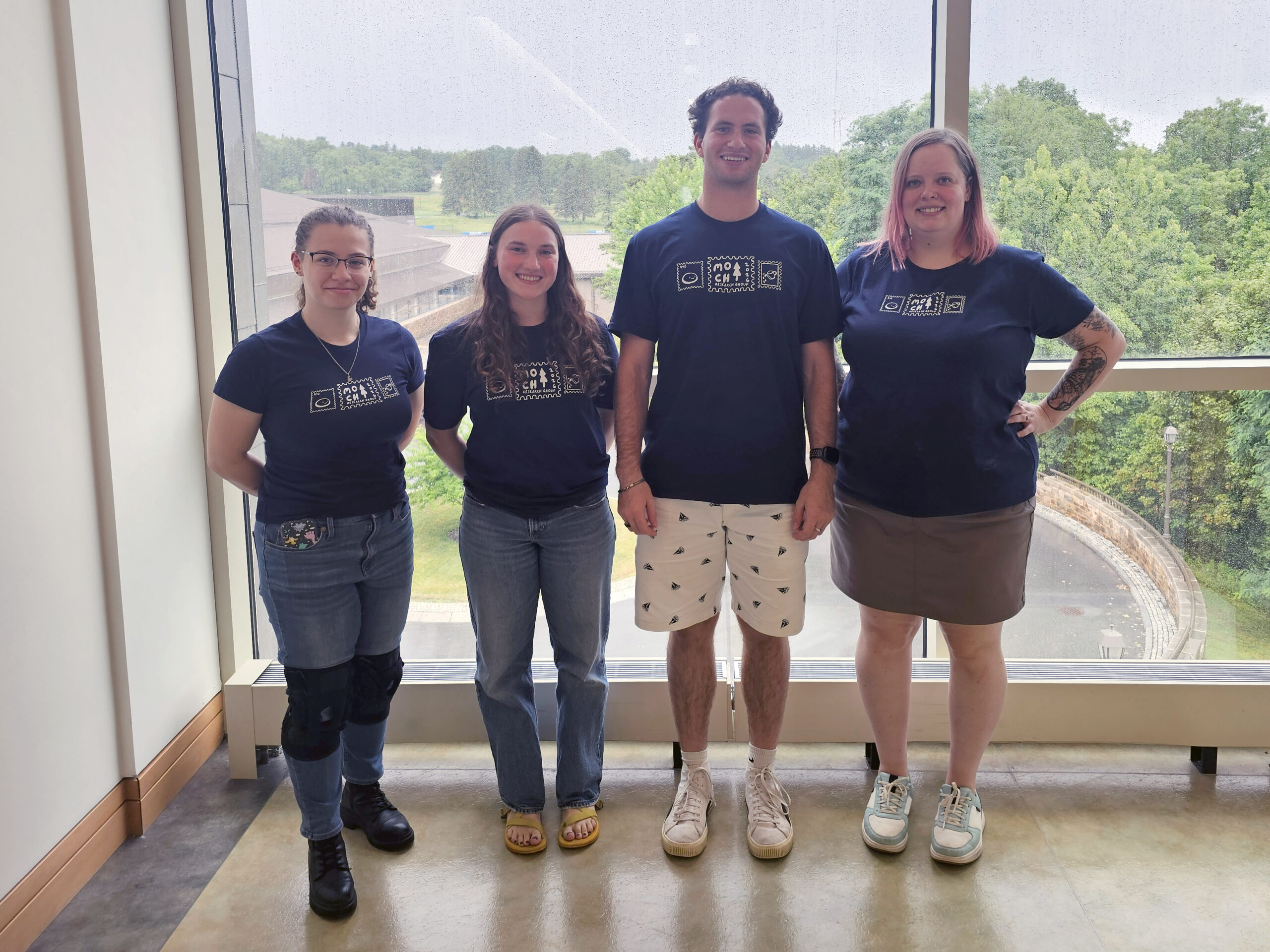 Four people are standing in front of a window wearing matching navy t shirts. From left to right, Madeline Brogen, Gwen Sawicki, Royce Carol, and Prof. Morrison-Smith.