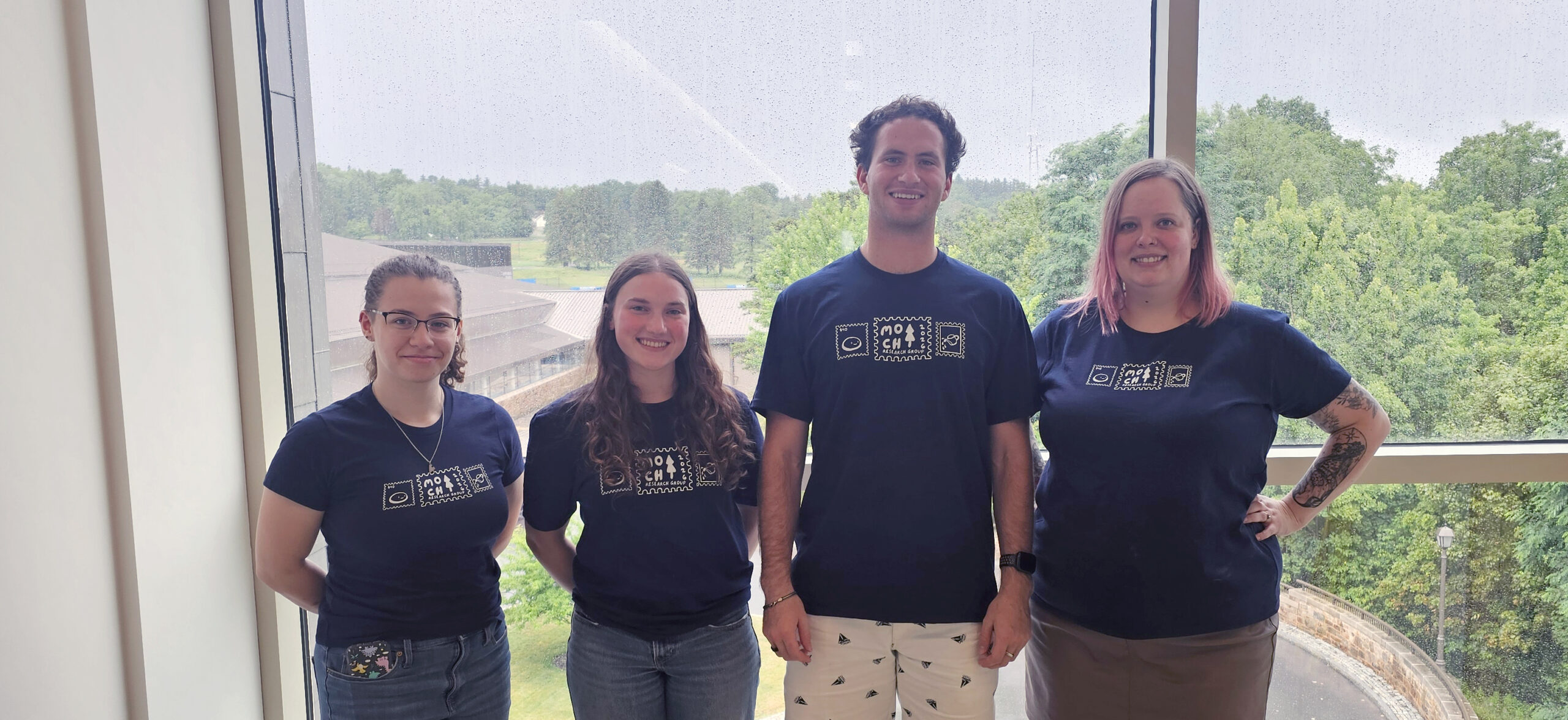 Madeline Brogen, Gwen Sawicki, Royce Carol, and Prof. Morrison-Smith stand in front of a floor length window while wearing matching navy t shirts